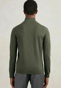 Donkergroen gebreide trui met een hoge kraag, lange mouwen en een aansluitend ontwerp. Soepele textuur met minimale naden, gecombineerd met grijze broeken.