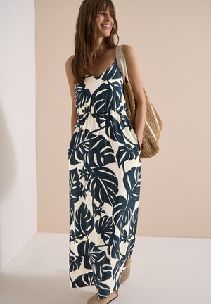 Femme souriante debout, portant une robe maxi blanche sans manches avec de grands motifs de feuilles bleu foncé et un sac à bandoulière tressé.