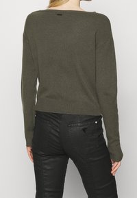 Zielonooliwkowy sweter dzianinowy z szerokim dekoltem i ściągaczem u dołu, zestawiony z czarnymi skórzanymi spodniami z tylnymi kieszeniami i detalem z logo.