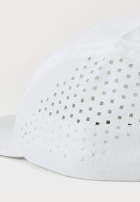 White cap made of breathable fabric featuring a textured pattern of small square cutouts, with a curved brim and an adjustable back.