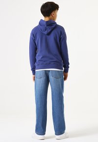 Sudadera azul con bolsillo canguro y cordón, combinada con jeans de pierna ancha azul claro y zapatillas blancas, vista desde la parte posterior.