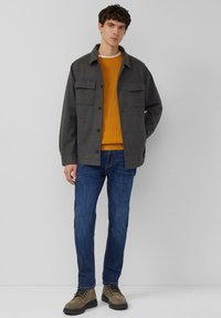 Młody mężczyzna ubrany w szarą zapinaną na guziki kurtkę na swetrze w kolorze musztardowym, niebieskie dżinsy i taupe zamszowe buty, stojący na tle jednolitego szarego tła.