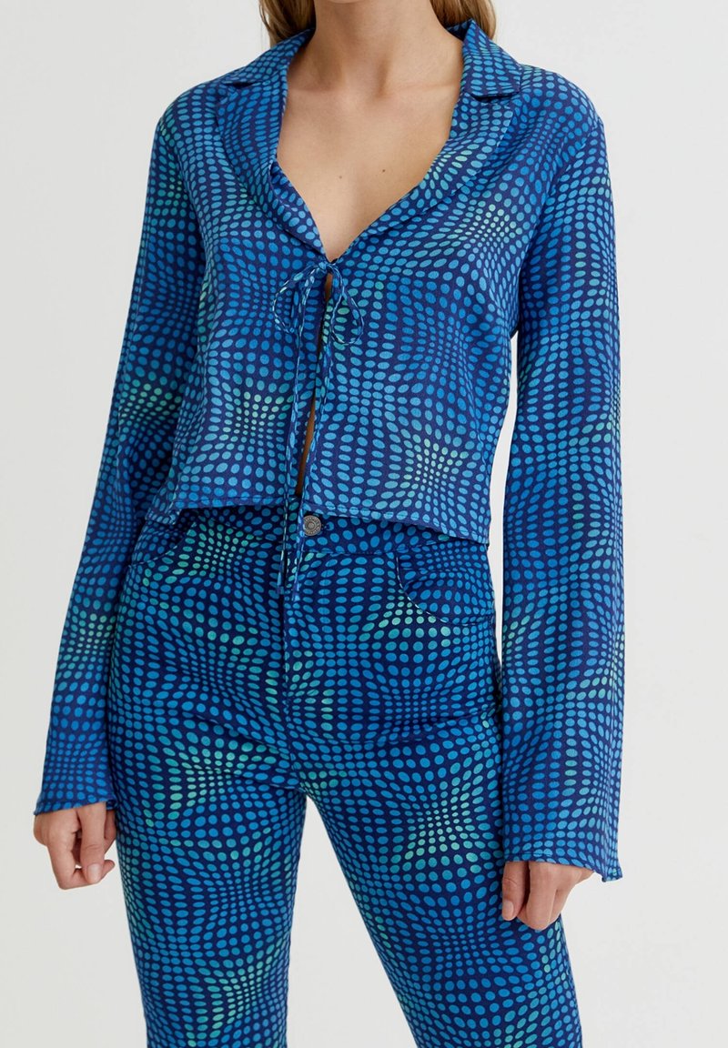 Femme portant un haut à manches longues bleu et un pantalon avec un motif circulaire à pois dans des nuances plus claires de bleu et de vert.