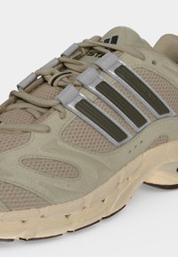 Μπεζ αθλητικό παπούτσι Adidas με λεπτομέρειες από δίχτυ και σουέτ, μαύρες ρίγες στο πλάι, μπεζ κορδόνια και υφασμένη σόλα σε ανοιχτό φόντο.