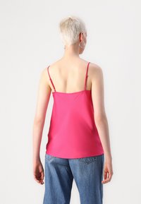 Fuchsia cami med tynne justerbare stropper, laget av et glatt stoff. Har en avslappet passform og en avrundet nederkant. Kombinert med denimshorts.