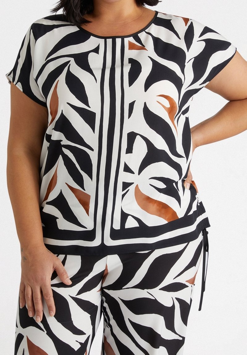Femme portant un haut manches courtes à motif abstrait de feuilles en noir, blanc et marron et pantalon assorti, main posée sur la hanche.