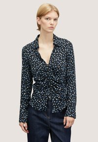 Čern á dlouhá rukávová košile s světle modrými oválnými vzory, na přední straně s nařasením a s límečkem. Spárováno s tmavými džíny.