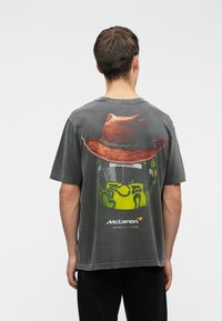 Graues Baumwoll-T-Shirt mit einem großen Grafikdesign eines braunen Huts auf der Rückseite, ausgestattet mit einem grünen und schwarzen Helmdesign sowie dem Text "McLaren Formel 1 Team".