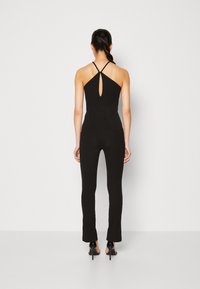 Femme portant une combinaison noire à col licou avec un détail en trou de serrure dans le dos et des sandales à talons hauts noires, debout devant un fond clair uni.