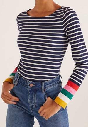 Marineblauw-wit gestreept shirt met lange mouwen en kleurrijke gestreepte manchetten, gecombineerd met een blauwe spijkerbroek met hoge taille. Handen rusten in de zakken.