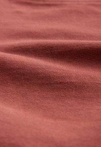 Mäkká tkanina s jemnou textúrou v tlmenej červeno-hnedej farbe s jemnými záhybmi a detailmi tkania viditeľnými na detailnom zábere.