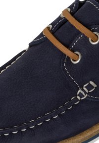 Chaussure en cuir bleu marine avec lacets en cuir marron, coutures blanches et œillets métalliques, montrant un savoir-faire détaillé et une texture.