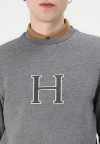 Grauer Sweatshirt mit hohem Kragen. Auf der Brust befindet sich ein auffälliger, gestickter Buchstabe "H" in Weiß. Weiche Textur.