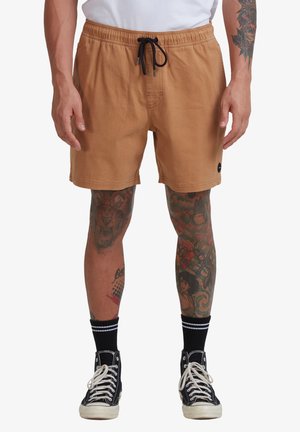 Brown swim shorts with an elastic waistband and black drawstring, side pockets, and a small logo patch near the hem. Black sneakers with white laces.