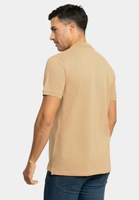 Camisa polo beige con cuello clásico y mangas cortas, hecha de una tela suave. Diseño ajustado combinado con unos vaqueros de denim azul oscuro.