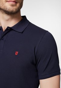 Granatowa koszulka polo wykonana z teksturowanej tkaniny, z klasycznym kołnierzykiem i małym, czerwonym haftowanym logo na piersi.