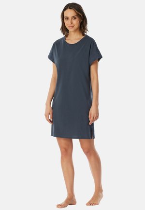 Woman with shoulder-length brown hair wearing a short-sleeve navy blue knee-length dress, standing barefoot on a white background.