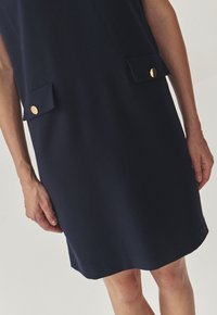 Navy blue dress made of soft fabric, featuring two front pockets with gold buttons and a simple, straight cut design.
