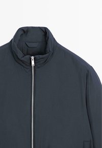 Granatowa marynarka z wysokim kołnierzem, zamkiem błyskawicznym z przodu i minimalistycznym designem. Wykonana z lekkiego materiału o gładkiej fakturze, bez widocznych wzorów.