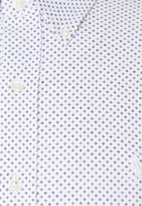 Bílá košile s knoflíky a malým modrým diamantovým vzorem a slonovinovými knoflíky, zobrazená s límcem a částečnou oblastí hrudníku.