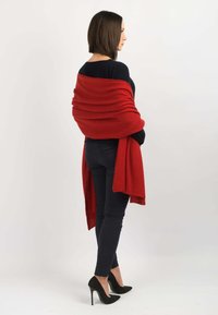 Rød tørklæde lavet af blødt stof, der hænger omkring skuldrene med løse ender. Båret over en mørk sweater og parret med sorte tætsiddende bukser og højhælede sko.