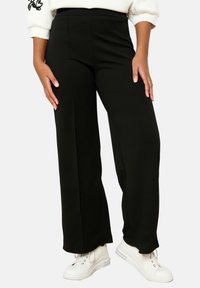 Melni platās kājas bikses, izgatavotas no gludas auduma, ar augstu jostasvietu un bez redzamām aizdarēm. Apvienotas ar baltām sporta apavām.