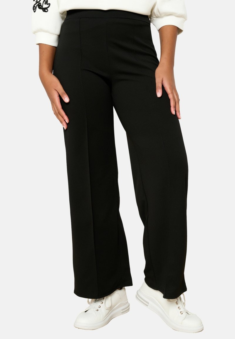 Melni platās kājas bikses, izgatavotas no gludas auduma, ar augstu jostasvietu un bez redzamām aizdarēm. Apvienotas ar baltām sporta apavām.