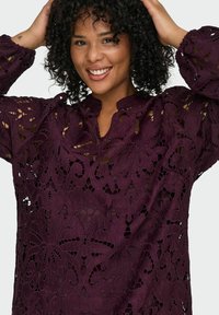 Burgundijska čipkana bluza s složenim cvjetnim izrezima, mandarinskim ovratnikom i dugim rukavima. Teksturirani uzorak povećava vizualne detalje.