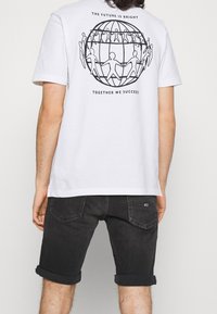 T-shirt blanc en coton avec un graphisme noir représentant un globe et des silhouettes se tenant la main au dos. Le texte "L'AVENIR EST BRILLANT" est inscrit au-dessus.