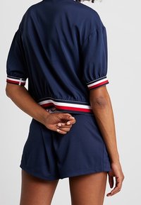 Marinblå kortærmet jakke med en cropped design, der har stribede detaljer i rød og hvid ved ærmerne og taljen, parret med matchende shorts.