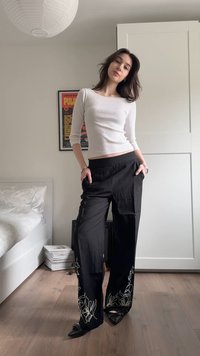 Femme debout à l'intérieur portant un haut blanc à manches longues, un pantalon noir à jambes larges avec une broderie blanche, et des chaussures noires à bouts pointus.