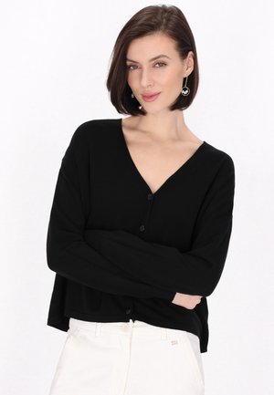Kvinde med kort mørkt hår iført en sort knappet sweater og hvide bukser står med armene krydset mod en ensfarvet hvid baggrund.