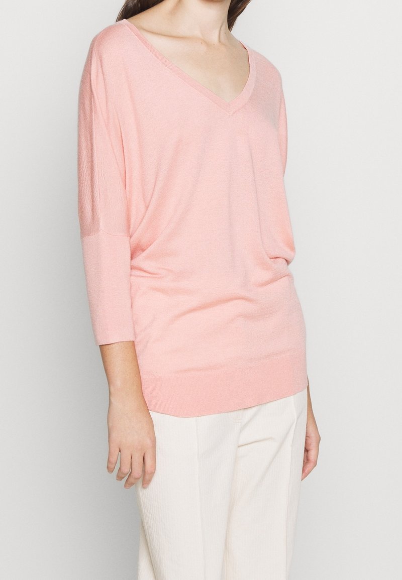 Femme portant un pull ample rose clair à col en V avec des manches trois-quarts et un pantalon blanc sur un fond uni.