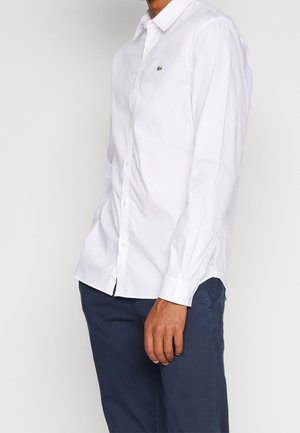 Homme portant une chemise blanche cintrée avec boutons, avec un petit logo vert sur la poitrine et un pantalon bleu foncé sur un fond uni.