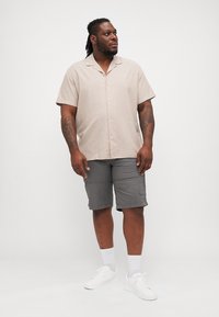 Camicia beige chiaro a maniche corte con bottoni, abbinata a pantaloni cargo grigi. Il modello indossa sneakers bianche e calzini bianchi di lunghezza media.