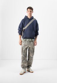 Un tânăr stă cu spatele la un fundal simplu, purtând un hanorac bleumarin, o cămașă verde în straturi, pantaloni cargo cu camuflaj și adidași, având o geantă crossbody.