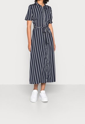 Vrouw draagt een midi-blousejurk met verticale strepen in marineblauw en wit, korte mouwen, knopen aan de voorkant, tailleband en witte platform sneakers.
