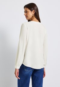 Femme aux longs cheveux bruns portant des boucles d'oreilles en or, un chemisier blanc à manches longues et un jean bleu, de dos et regardant sur le côté.