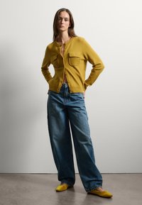 Cardigan giallo senape con due tasche sul petto, chiusura a bottoni, abbinato a jeans blu a vestibilità ampia e ballerine gialle.