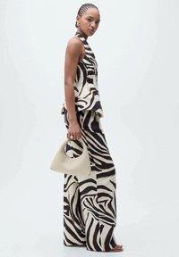 Ärmelloses Oberteil mit Zebra-Muster und weit geschnittene Hosen in Beige und Schwarz. Hält eine kleine, strukturierte, helle Handtasche. Glattes Material.