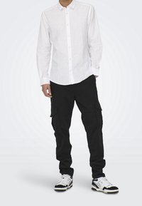 Camisa blanca de manga larga con botones, combinada con pantalones cargo negros con bolsillos laterales y zapatillas deportivas en blanco y negro.