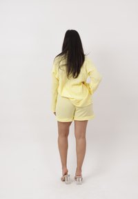 Mujer con cabello largo y oscuro de espaldas, lleva una camisa amarilla suelta, pantalones cortos a juego y sandalias transparentes con tacón sobre un fondo liso.