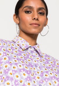 Blusa floral em roxo claro com margaridas brancas e detalhes em laranja. Possui colarinho, fecho de botão e mangas curtas. Textura suave.