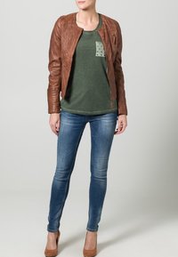 Hnědá kožená bomber bunda s texturovaným povrchem, zelené tričko s kapsou a cvočky, modré slim fit džíny a hnědé podpatky.
