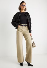 Γυναίκα με μαύρο bomber jacket, μαύρο τοπ, μπεζ παντελόνι φαρδύ, μαύρες μυτερές γόβες, μπλε γυαλιά, κρατώντας μικρό τσαντάκι από δέρμα φιδιού.