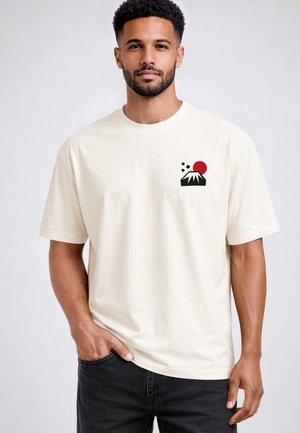 Mann mit kurzem lockigem Haar und Bart trägt ein weißes T-Shirt mit kleinem schwarzem und rotem Vulkan-Design auf der Brust, steht mit einer Hand in der Tasche.
