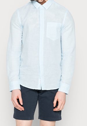 Homme portant une chemise bleu clair à manches longues avec une poche poitrine et un short bleu marine foncé sur un fond uni.