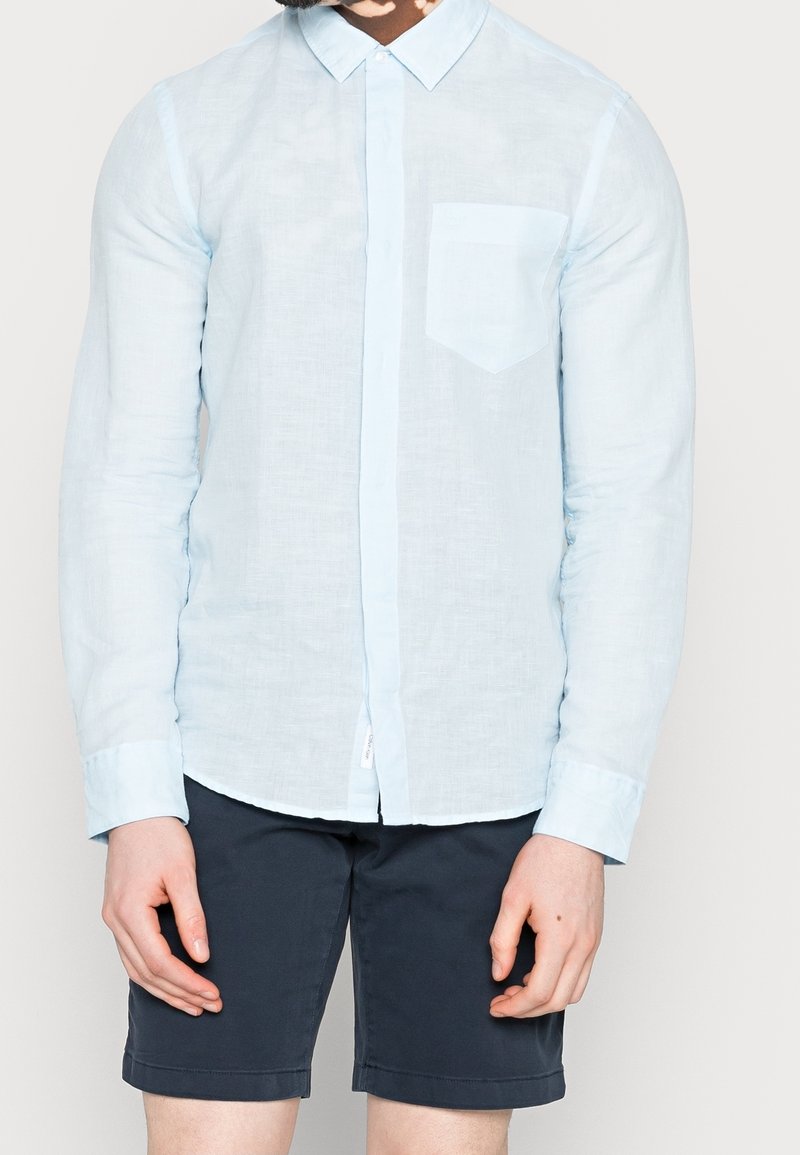 Homme portant une chemise bleu clair à manches longues avec une poche poitrine et un short bleu marine foncé sur un fond uni.