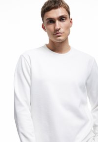 Hvid ribbet langærmet skjorte med rund hals, glat tekstur og tætsiddende design. Stoffet fremstår blødt og let.