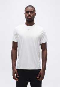 Άνδρας που στέκεται εξερχόμενος προς τα εμπρός φορώντας λευκό αθλητικό μπλουζάκι με κοντά μανίκια, ανοιχτοπράσινες πλαϊνές λωρίδες και μαύρο σορτς, μπροστά σε λευκό φόντο.